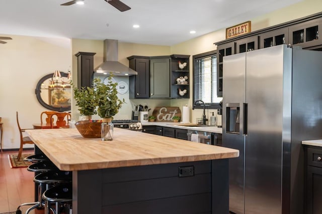 kitchen with wall chimney exhaust hood, stainless steel fridge with ice dispenser, butcher block countertops, a kitchen island, and hardwood / wood-style flooring