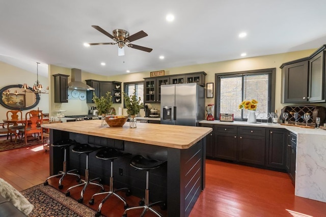 kitchen with wall chimney exhaust hood, stainless steel fridge with ice dispenser, dark hardwood / wood-style flooring, wooden counters, and a kitchen island