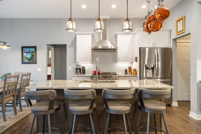 kitchen with stainless steel refrigerator with ice dispenser, wall chimney range hood, decorative light fixtures, white cabinetry, and an island with sink