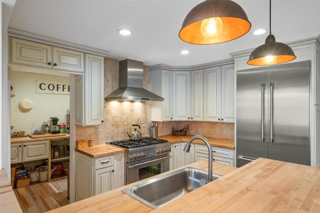 kitchen featuring sink, wall chimney exhaust hood, premium appliances, wooden counters, and decorative light fixtures