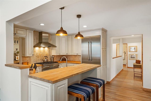 kitchen featuring high end appliances, backsplash, wooden counters, wall chimney range hood, and kitchen peninsula