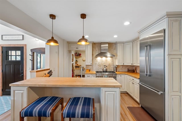 kitchen with wood counters, stove, a kitchen breakfast bar, wall chimney range hood, and stainless steel built in fridge