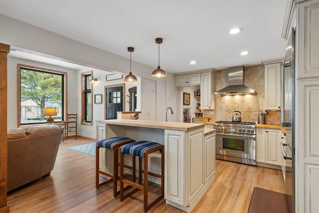 kitchen with decorative backsplash, wall chimney range hood, a center island with sink, high end stainless steel range, and hanging light fixtures