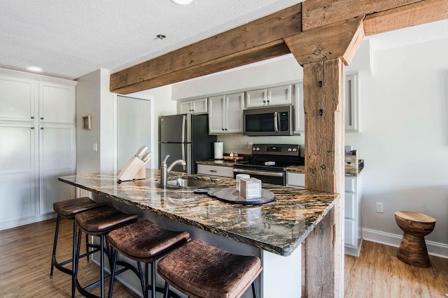 kitchen featuring a kitchen bar, stainless steel appliances, beamed ceiling, dark stone countertops, and light wood-type flooring