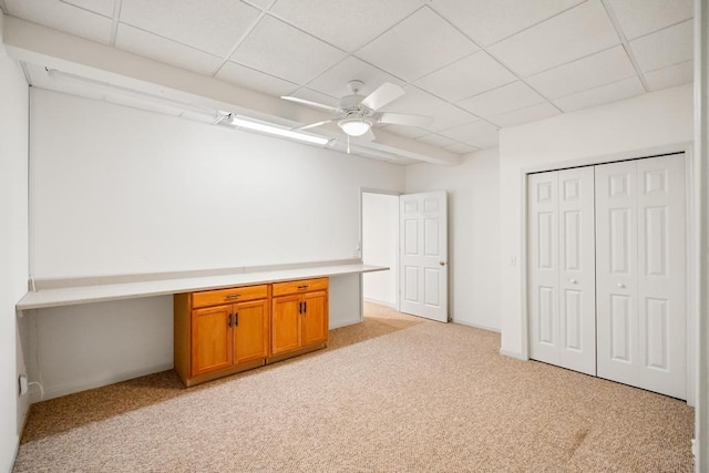 basement featuring light carpet, built in desk, ceiling fan, and a paneled ceiling