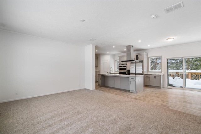 kitchen with gray cabinetry, island range hood, light carpet, stainless steel appliances, and a kitchen island with sink