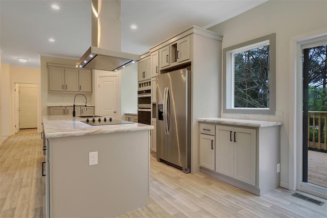 kitchen with light stone counters, appliances with stainless steel finishes, an island with sink, island exhaust hood, and light hardwood / wood-style floors