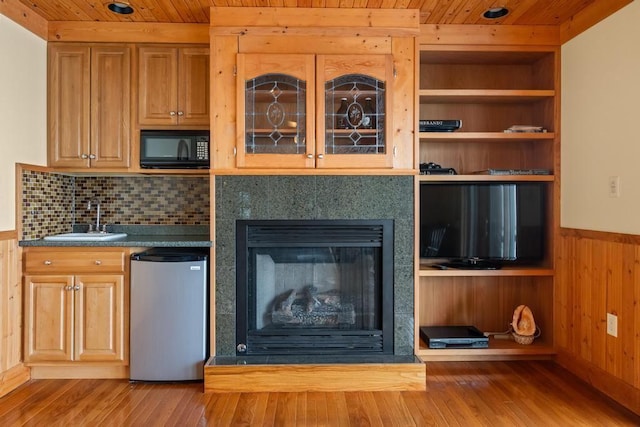 interior space featuring a wainscoted wall, light wood-style flooring, a glass covered fireplace, a sink, and wooden ceiling