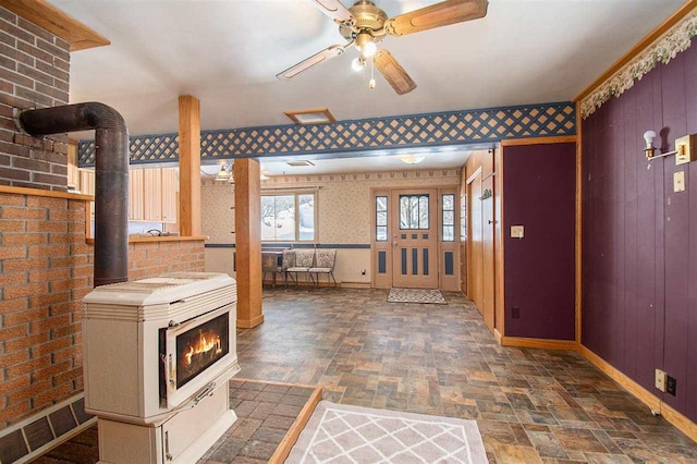 interior space featuring a wood stove, stone finish flooring, ceiling fan, baseboards, and wallpapered walls