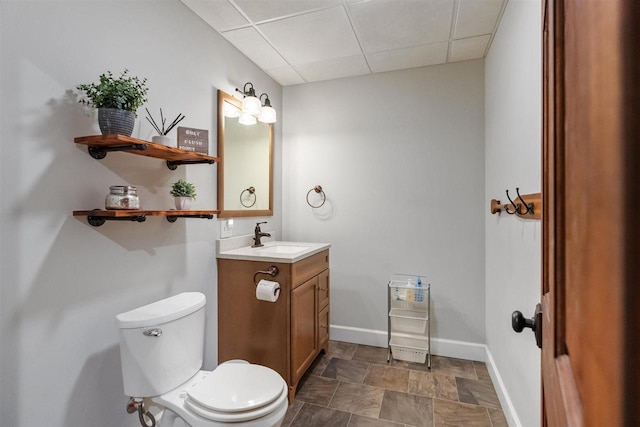 bathroom featuring baseboards, toilet, stone finish flooring, vanity, and a paneled ceiling