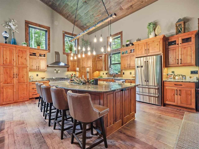 kitchen with high vaulted ceiling, high end fridge, light wood-type flooring, wall chimney exhaust hood, and plenty of natural light