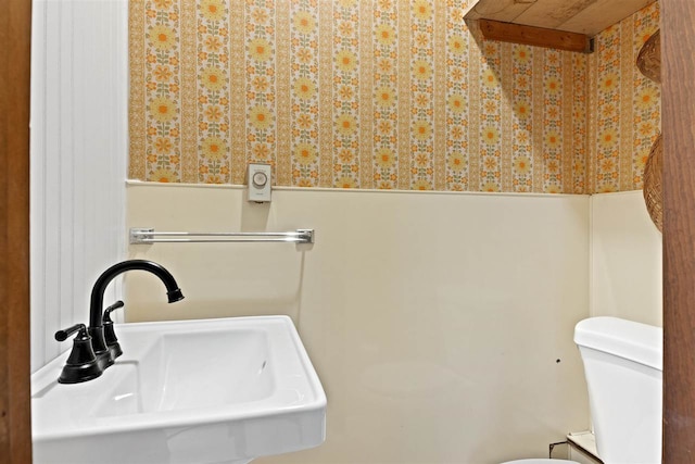 half bath featuring toilet, wallpapered walls, a sink, and a wainscoted wall