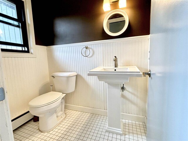 half bath with a baseboard radiator, wainscoting, tile patterned flooring, toilet, and baseboards