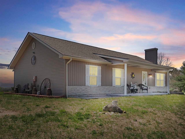 10300 Vista Ridge Dr, Alden, MI 49612 - back of property at dusk with a chimney, a lawn, board and batten siding, stone siding, and roof with shingles
