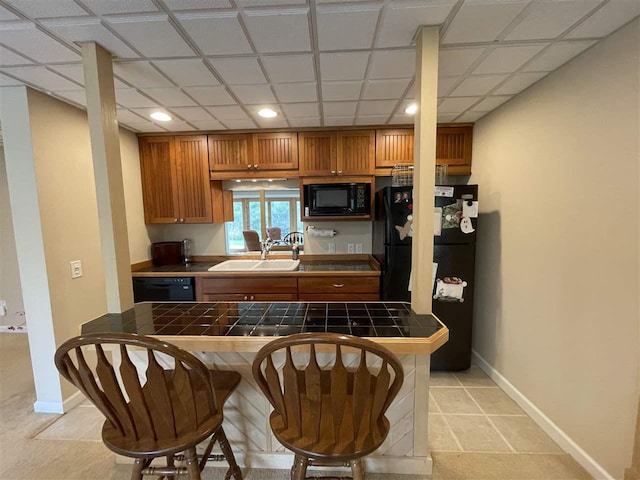 9030 Sugar Tree Ln, Charlevoix, MI 49720 - kitchen with tile counters, black appliances, brown cabinets, a kitchen breakfast bar, and baseboards