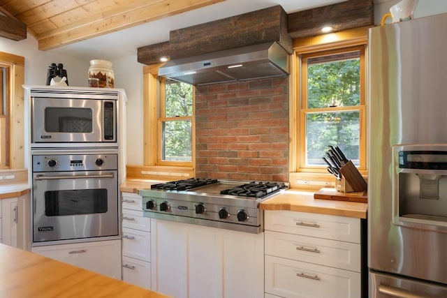 03675 Bc/Ej Rd, East Jordan, MI 49727 - kitchen with appliances with stainless steel finishes, butcher block counters, white cabinets, wood ceiling with exposed beams, and wall chimney range hood