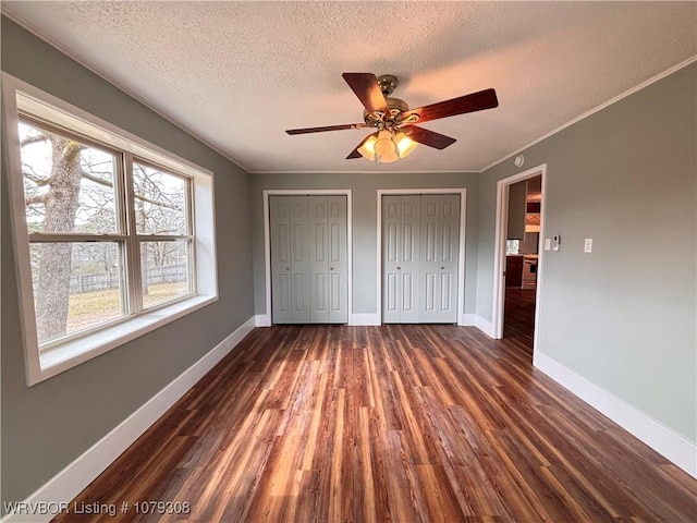 unfurnished bedroom featuring dark wood finished floors, two closets, ornamental molding, a textured ceiling, and baseboards