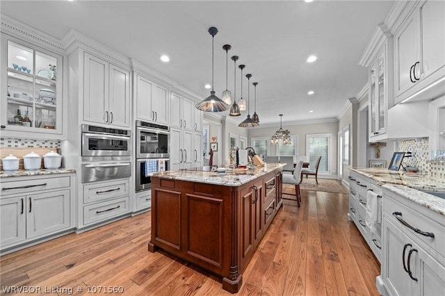 kitchen with pendant lighting, crown molding, double oven, an island with sink, and white cabinets