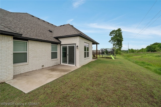rear view of property with a trampoline, a shingled roof, brick siding, a patio area, and a yard