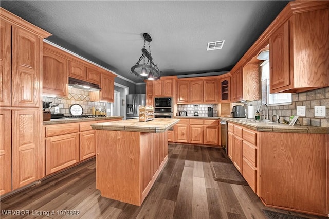 kitchen featuring a center island, under cabinet range hood, tile countertops, visible vents, and appliances with stainless steel finishes