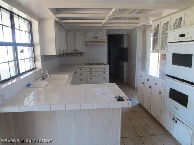 kitchen with kitchen peninsula, white double oven, sink, light tile patterned floors, and white cabinetry