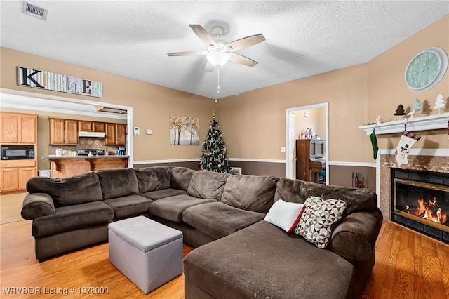 living room with a textured ceiling, light hardwood / wood-style flooring, ceiling fan, and a tiled fireplace