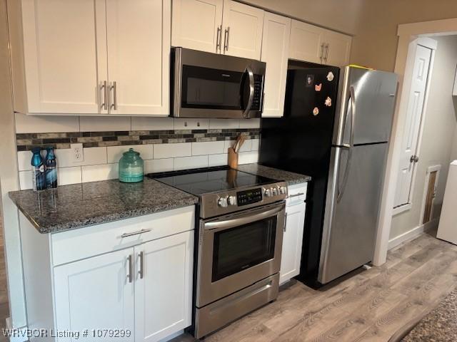 kitchen with white cabinets, dark stone counters, appliances with stainless steel finishes, light wood-type flooring, and backsplash