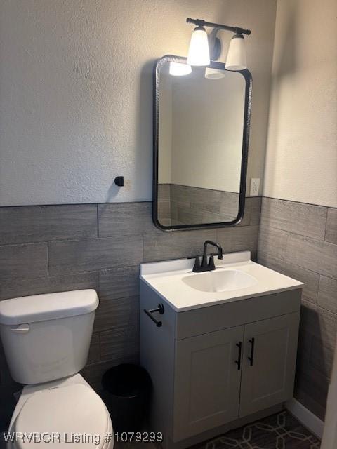 bathroom with a textured wall, toilet, vanity, tile walls, and wainscoting