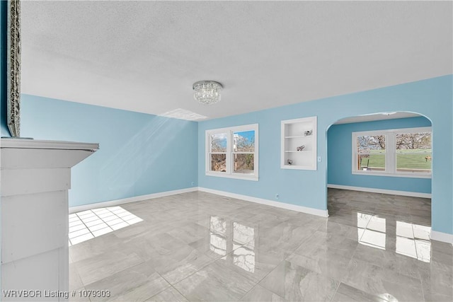 unfurnished living room featuring arched walkways, built in shelves, a textured ceiling, and baseboards