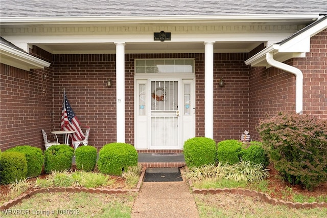 view of exterior entry with a porch, a shingled roof, and brick siding