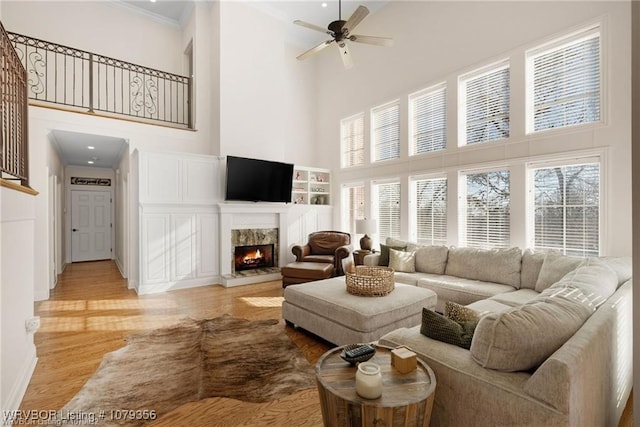 living area featuring a high ceiling, a premium fireplace, light wood-style floors, a ceiling fan, and ornamental molding