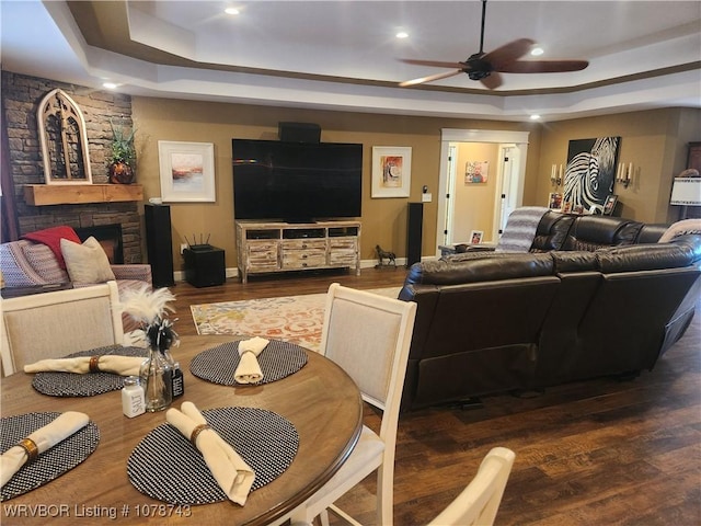 living room with dark hardwood / wood-style flooring, a stone fireplace, a raised ceiling, and ceiling fan