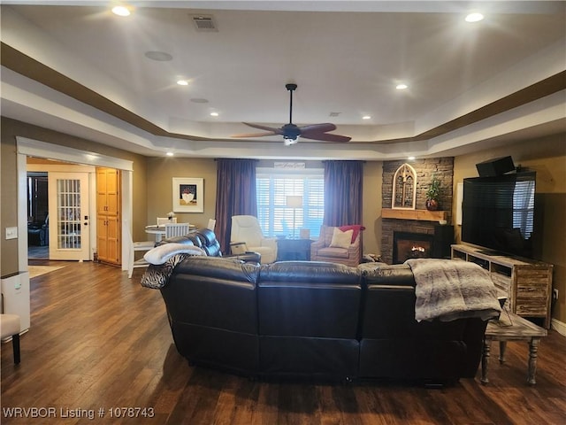 living room with dark wood-type flooring, ceiling fan, a stone fireplace, and a raised ceiling