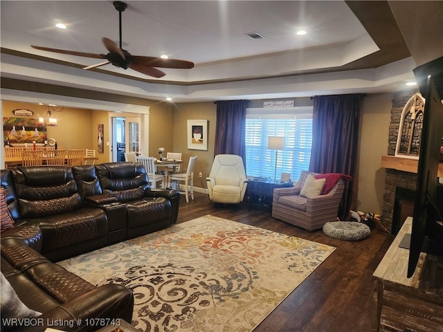 living room featuring dark hardwood / wood-style flooring, a stone fireplace, a raised ceiling, and ceiling fan