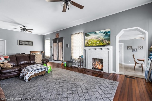 living room with ceiling fan, wood-type flooring, a brick fireplace, baseboards, and arched walkways