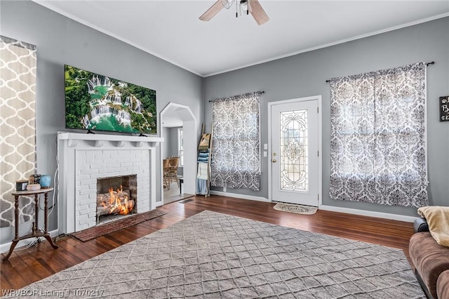 living room with wood finished floors, ceiling fan, a brick fireplace, and crown molding