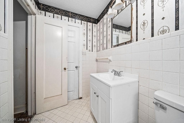 half bath with wallpapered walls, a textured ceiling, toilet, tile patterned floors, and a wainscoted wall