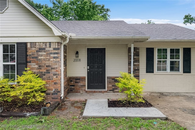 view of exterior entry featuring roof with shingles, brick siding, covered porch, and stone siding