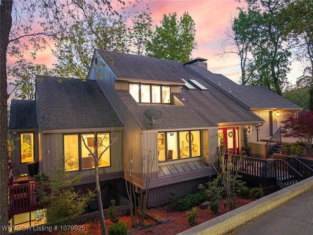 back of house at dusk featuring a chimney, a wooden deck, and roof with shingles