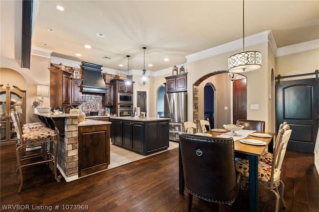 kitchen with appliances with stainless steel finishes, premium range hood, dark brown cabinets, pendant lighting, and an island with sink