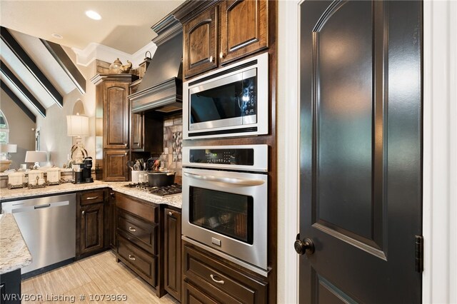 kitchen with lofted ceiling, premium range hood, appliances with stainless steel finishes, light stone counters, and dark brown cabinetry