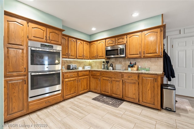 kitchen featuring brown cabinetry, light stone counters, stainless steel appliances, and backsplash