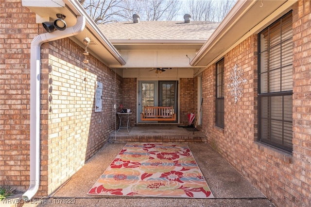 view of exterior entry with roof with shingles, brick siding, ceiling fan, and a patio