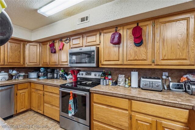 461510 E 1130th Rd, Sallisaw, OK 74955 - kitchen featuring appliances with stainless steel finishes, light tile patterned flooring, a textured ceiling, backsplash, and brown cabinetry