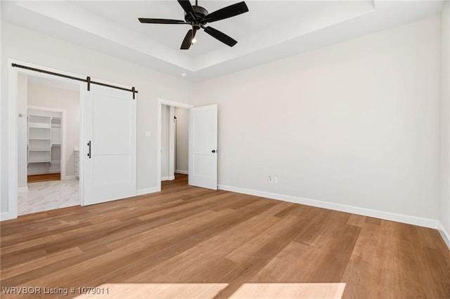 unfurnished bedroom with a spacious closet, ceiling fan, a tray ceiling, a barn door, and light hardwood / wood-style flooring