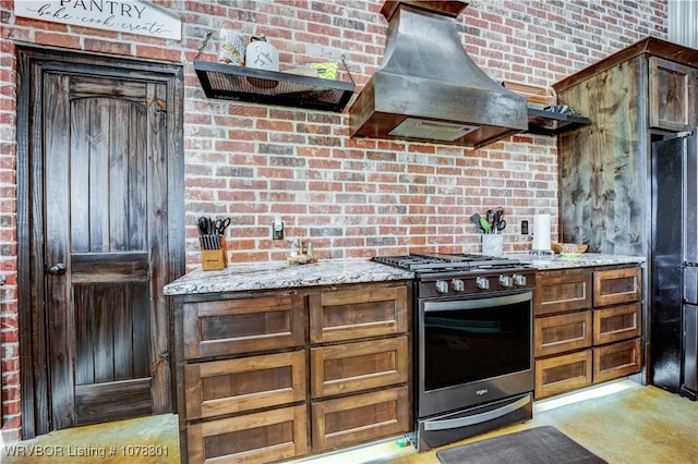 kitchen with wall chimney range hood, stainless steel range with gas cooktop, light stone counters, fridge, and dark brown cabinets