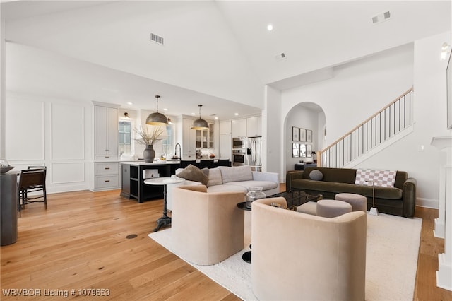 living room with visible vents, high vaulted ceiling, arched walkways, light wood-style floors, and a decorative wall