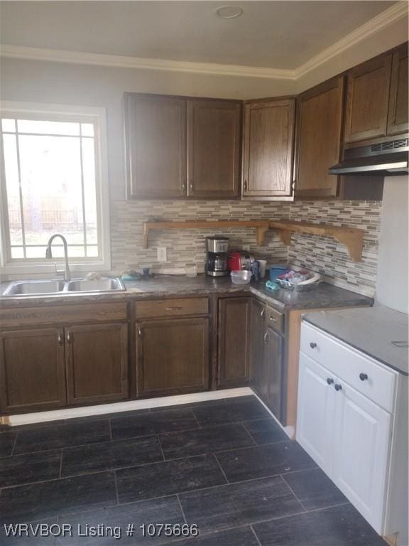 kitchen with under cabinet range hood, a sink, backsplash, dark countertops, and crown molding