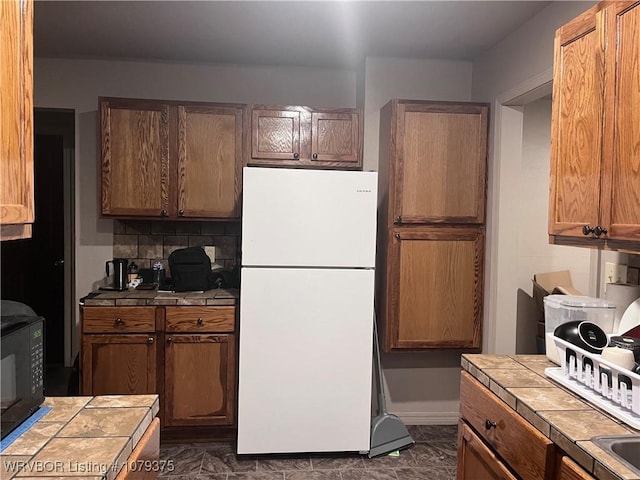 kitchen featuring black microwave, tile counters, decorative backsplash, brown cabinets, and freestanding refrigerator