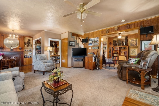 carpeted living room featuring a textured ceiling, built in shelves, wooden walls, and ceiling fan with notable chandelier